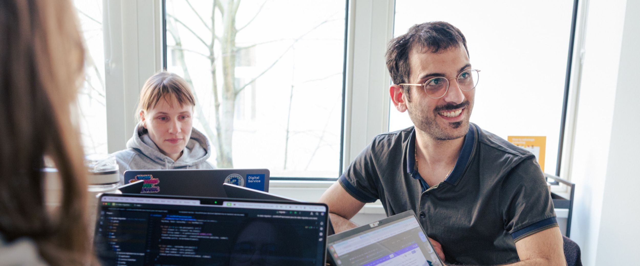 Three people are sitting in an office working on their laptops in front of them. Code can be seen on the screens. In the background is a window through which light falls.
