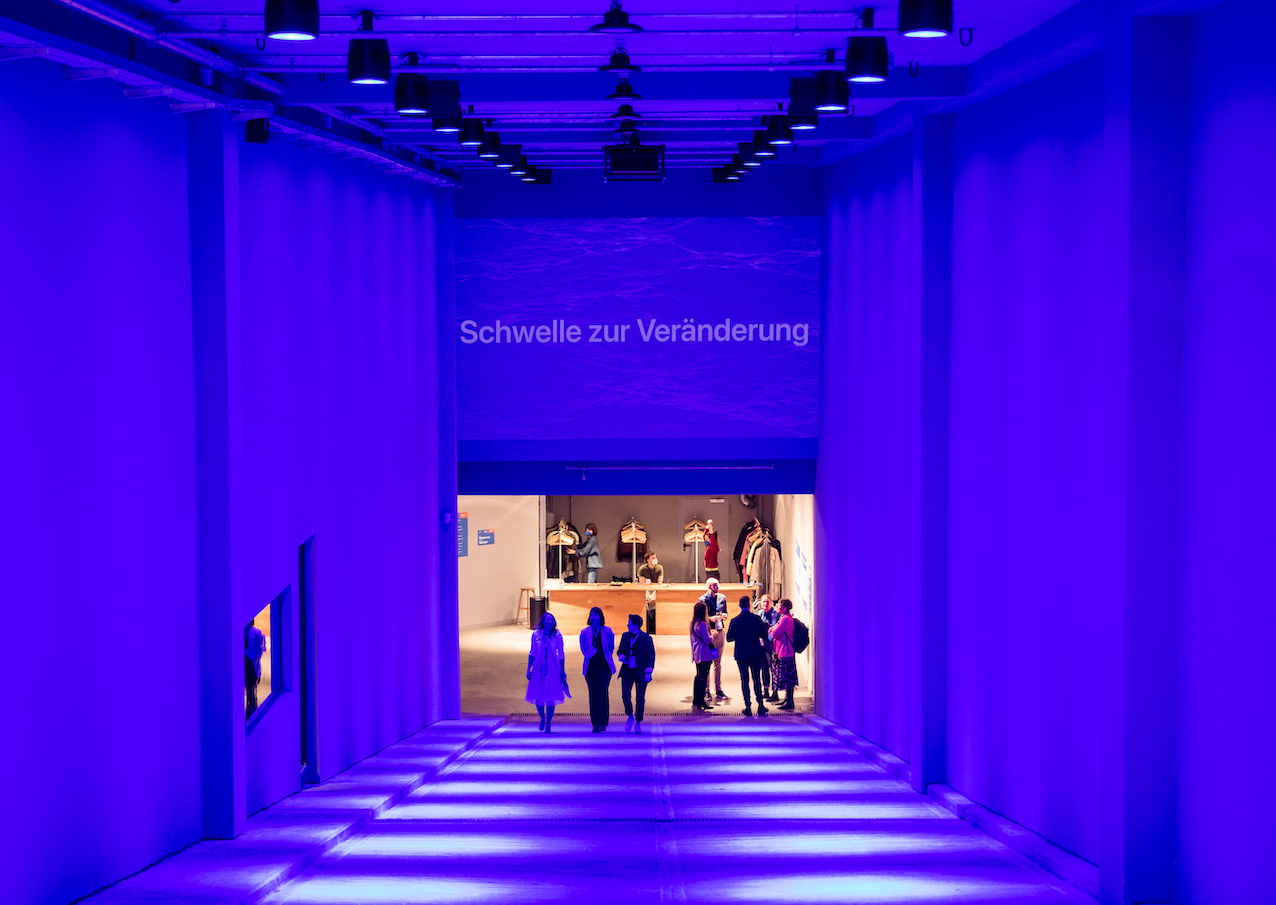 A group of people walk down a sloping, tunnel-like entrance with high walls toward a brightly lit dressing room; the entrance is shrouded in blue light; 