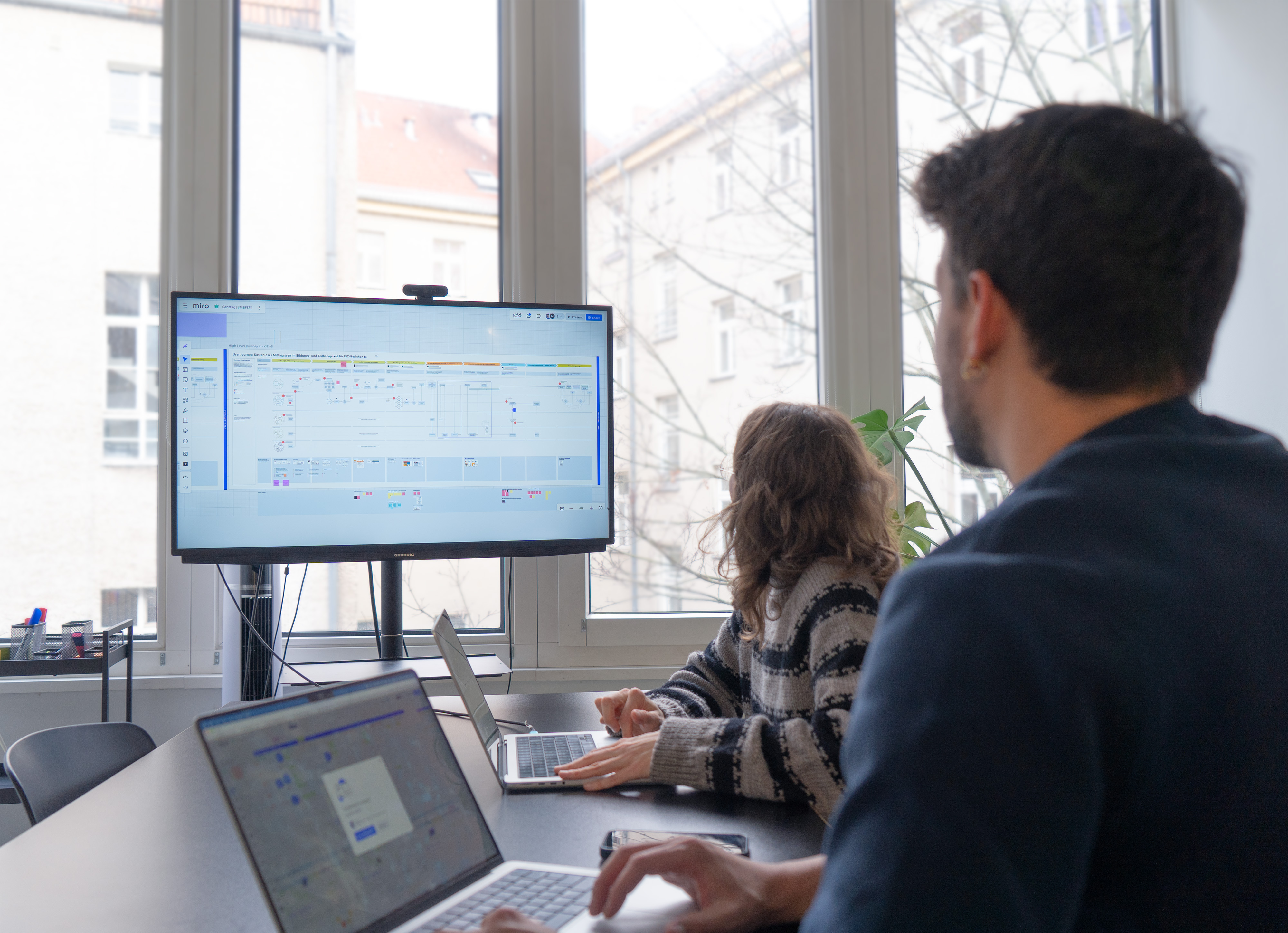 Zwei Personen sitzen in einem modernen Büroraum und schauen auf einen großen Bildschirm auf dem eine Grafik dargestellt wird. Vor Ihnen stehen geöffnete Laptops, im Hintergrund fällt Licht durch eine Fensterfront.