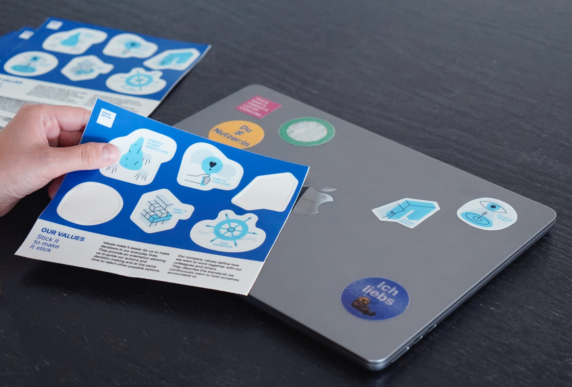 Eine Hand hält einen Bogen mit unterschiedlichen Stickern, darunter liegt ein Laptop auf dem bereits mehrere Sticker kleben.