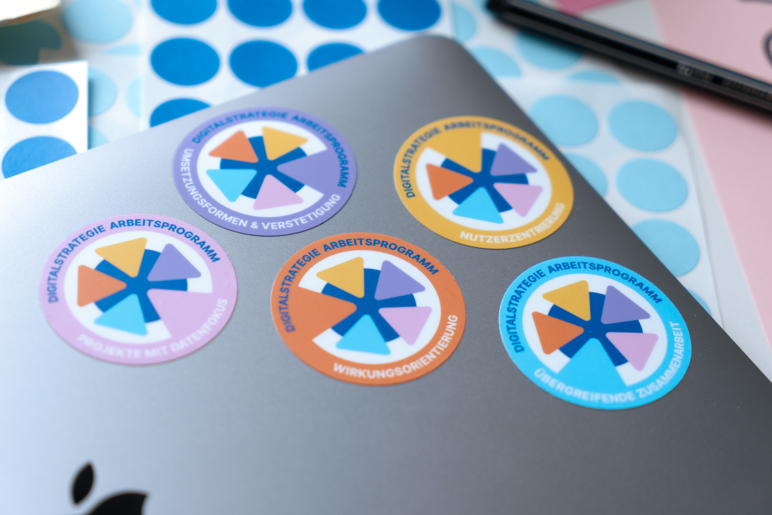 Auf einem Laptop sind fünf verschiedene Sticker in bunten Farben aufgeklebt, die für Teilnehmende des Arbeitsprogramms der Digitalstrategie bereitgestellt wurden