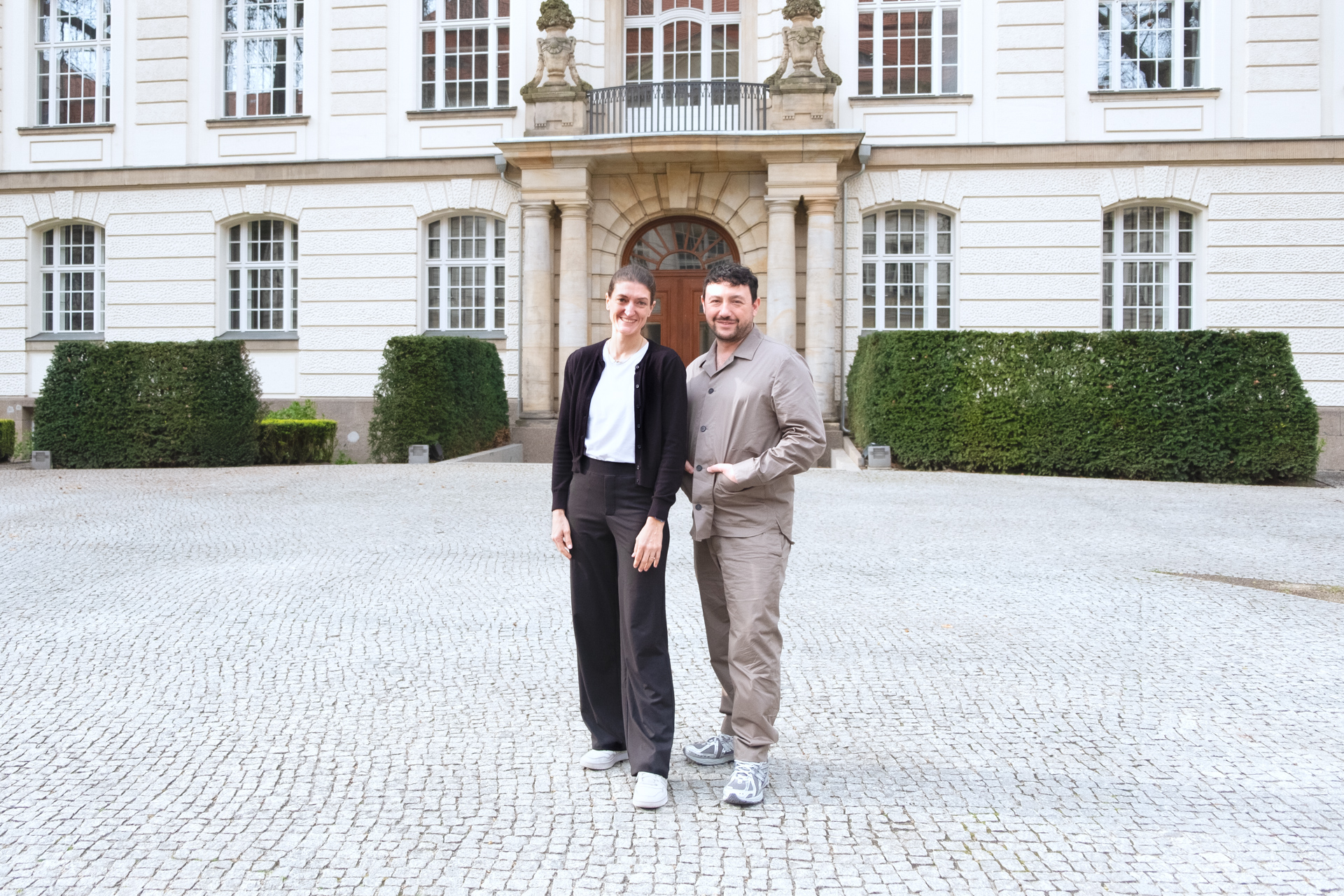 Julia und Önder stehen gemeinsam vor dem Eingang des BMWK Gebäudes, einem historischen Bau aus den Anfängen des 20. Jahrhunderts