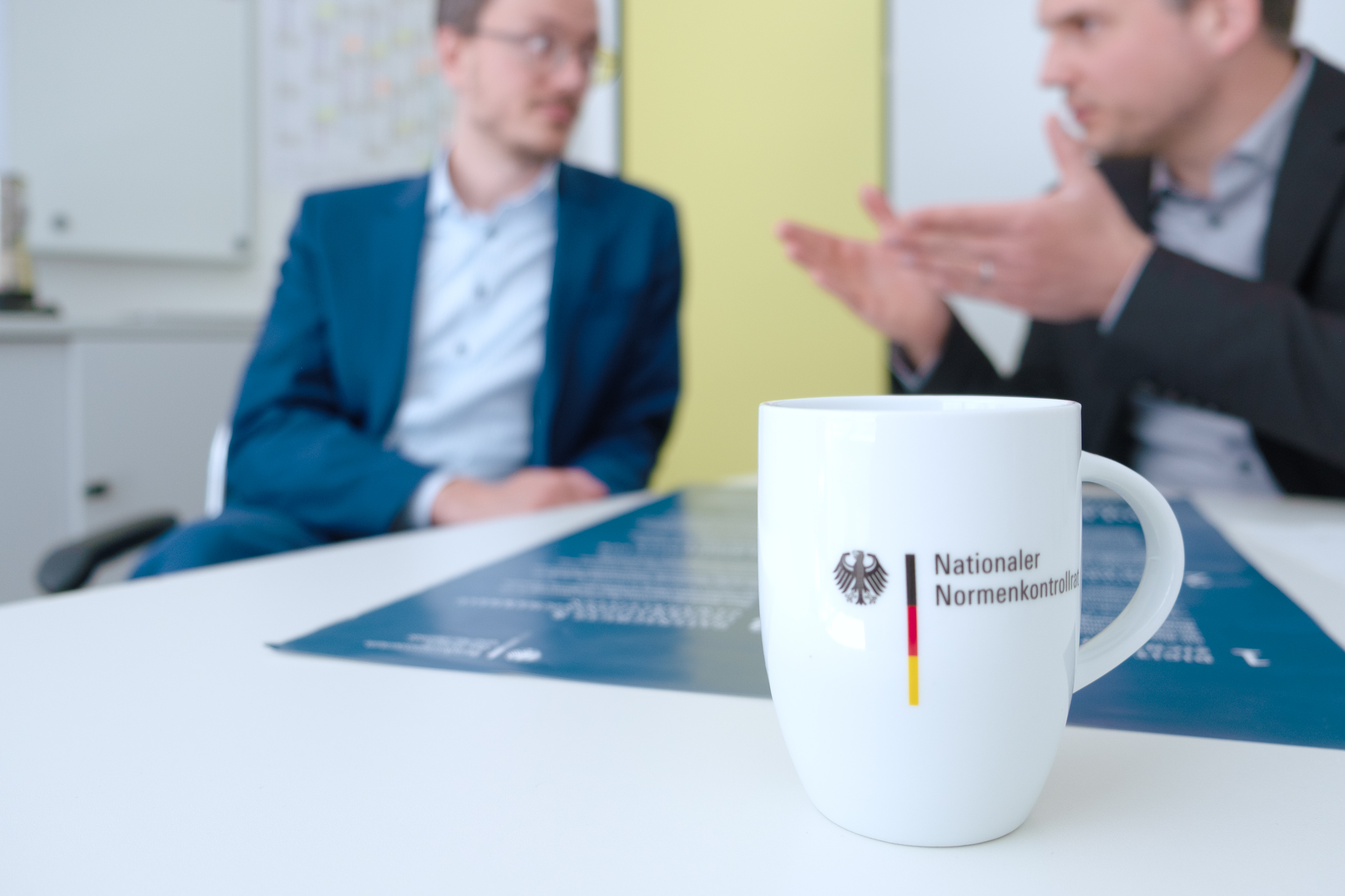 Im Vordergrund steht eine Tasse mit der Aufschrift „Nationaler Normenkontrollrat“. Im Hintergrund sind Patrick Eckner und Yannick Vogel im Gespräch zu erkennen.