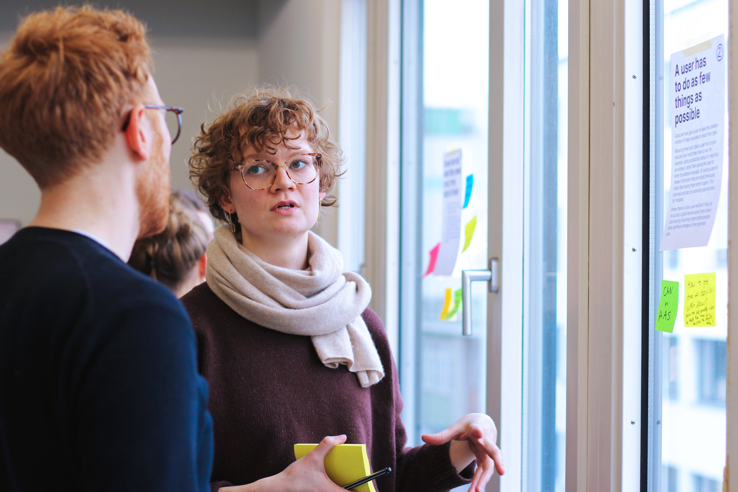 Zwei Personen unterhalten sich vor einem Fenster, an dem Notizzettel und kleine Poster kleben; auf einem im Fokus steht: „Ein Nutzender muss so wenig tun wie möglich.“