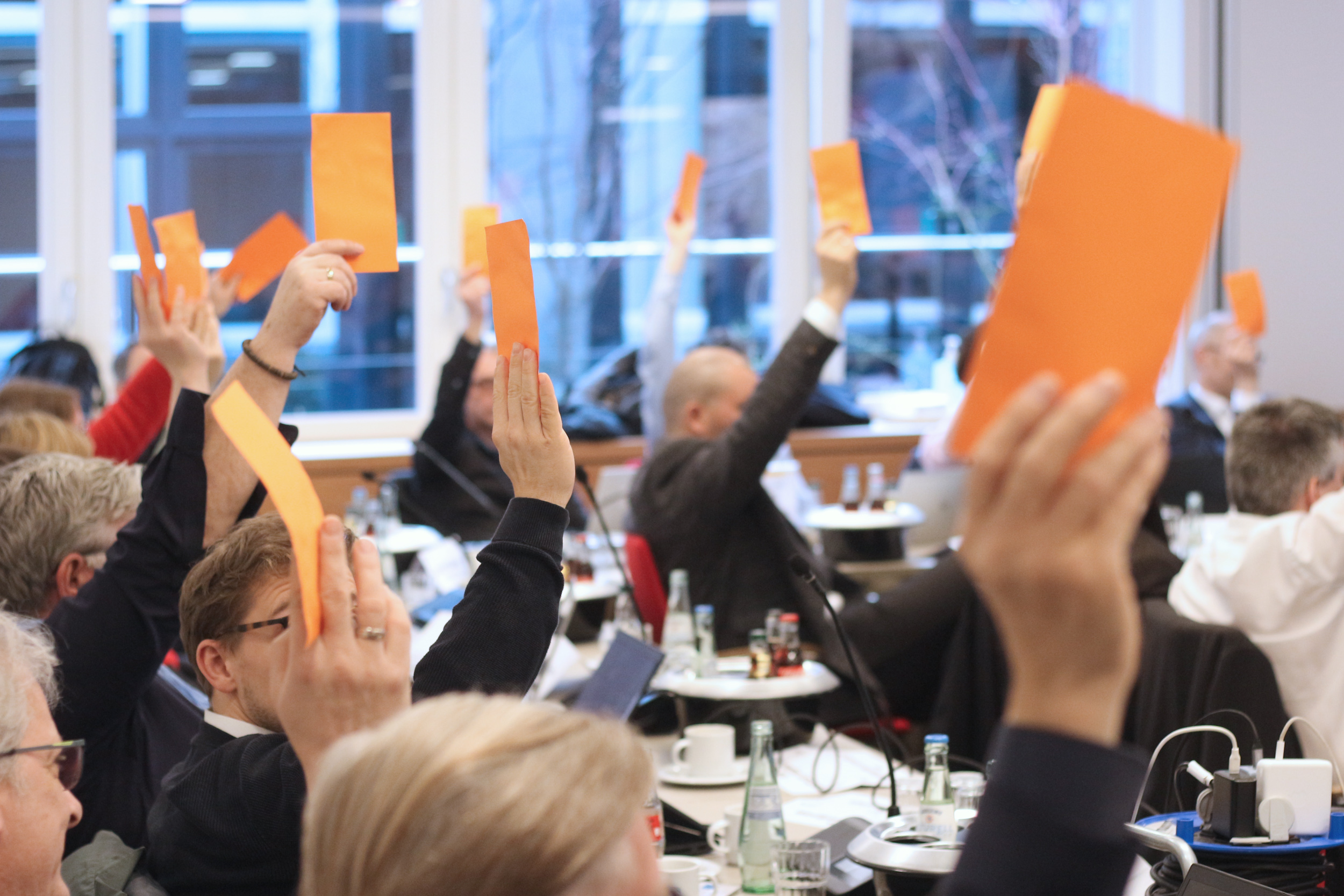 A group of people in a meeting room raise orange cards at the adoption of DIN SPEC 66336. The people sit at tables with drinks and documents while actively participating in the meeting.