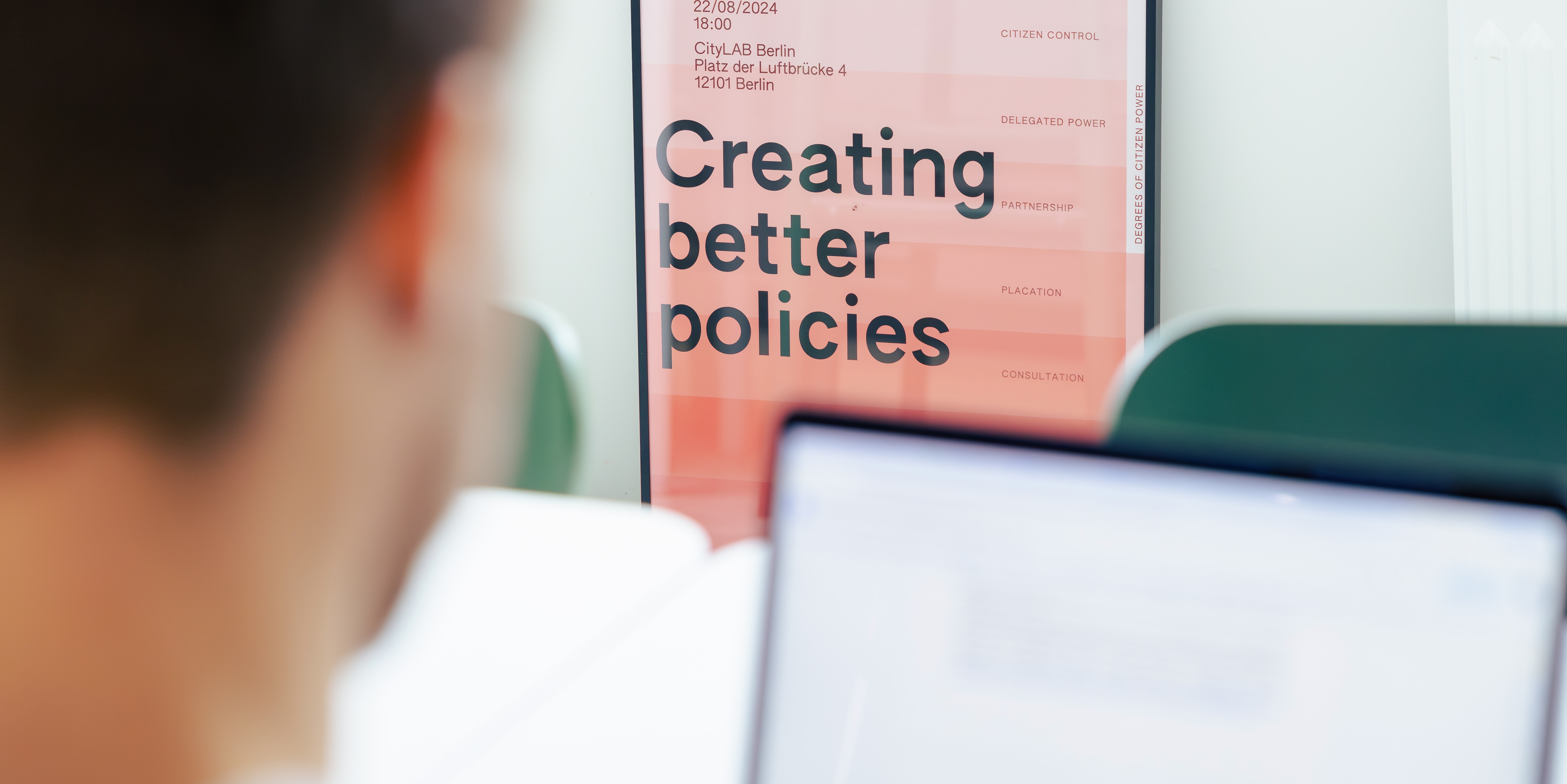 Auf einem Plakat steht „Creating better policies“. Vor dem Plakat ist die Silhouette einer Person, die am Laptop arbeitet, zu erkennen.