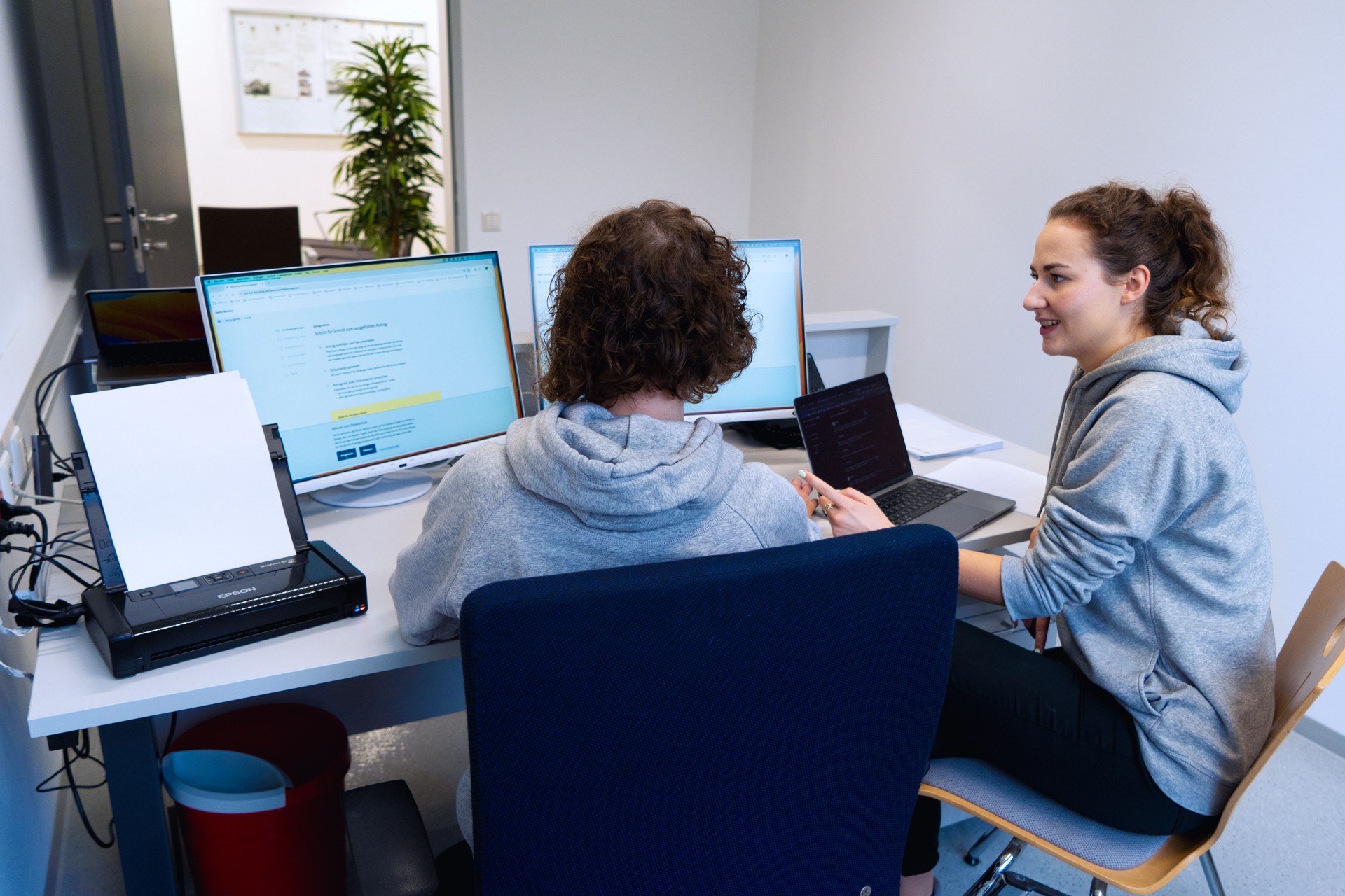 Zwei Personen sitzen an einem Schreibtisch mit mehreren Monitoren und unterhalten sich. Die Person links, deren Rückseite zu sehen ist, arbeitet an einem Computer. Die Person rechts, eine Frau im grauen Hoodie, hält einen Laptop und spricht. Ein Drucker und mehrere Kabel sind ebenfalls auf dem Schreibtisch sichtbar.