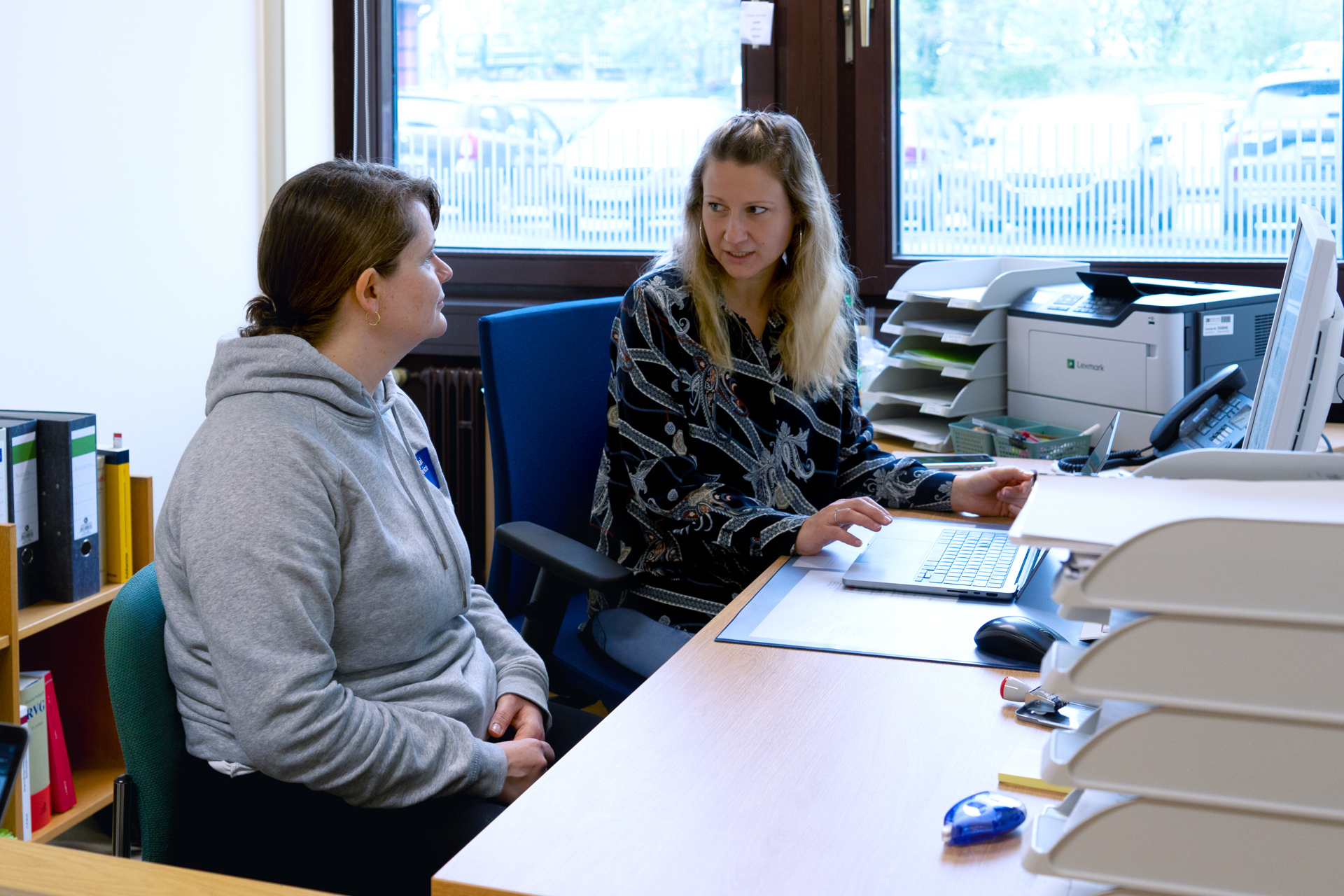 Zwei Frauen sitzen in einem Büro und unterhalten sich. Die Frau links trägt einen grauen Hoodie und sitzt mit verschränkten Händen. Die Frau rechts trägt ein gemustertes Oberteil und arbeitet an einem Laptop. Im Hintergrund sind ein Fenster, Aktenordner und Büromaterialien zu sehen.