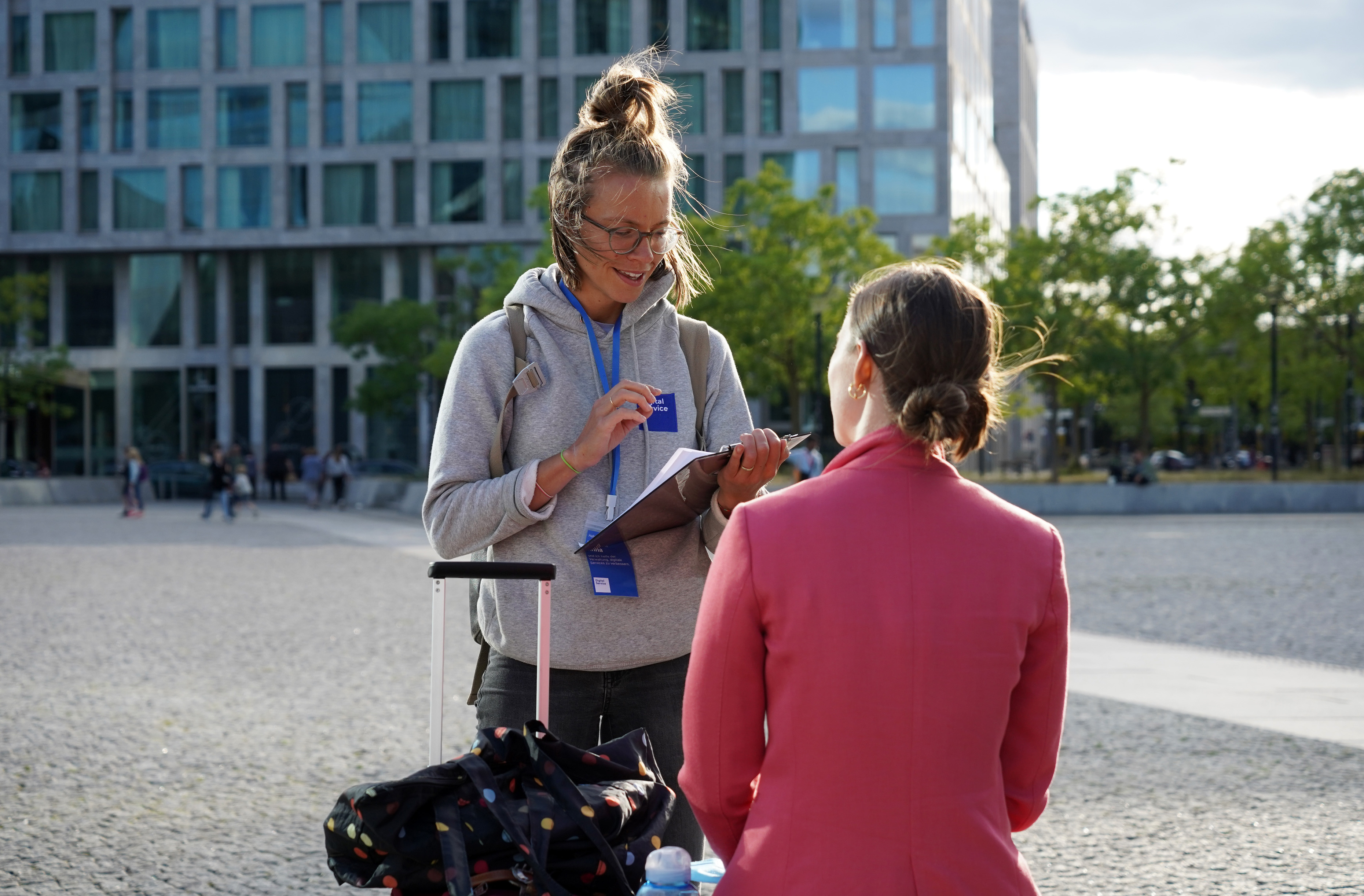 Two young women are talking to each other: one is standing, the other is sitting; the standing woman is holding a clipboard, wearing a hoodie with the DigitalService logo and a blue name tag, the sitting woman is wearing a pink blazer and gold earrings and is looking at them.