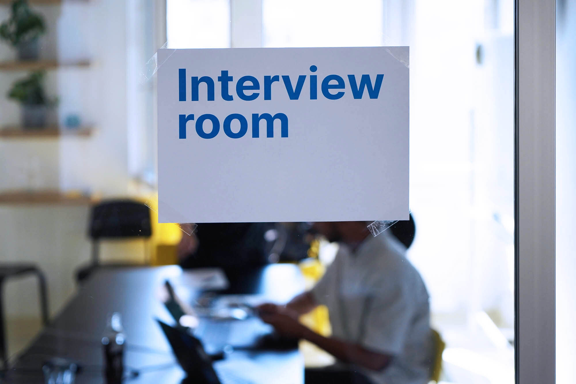 Ein Zettel an einer Glaswand, auf dem steht: “Interview room’. Dahinter sieht man unscharf 2 Menschen an einem Tisch sitzend.