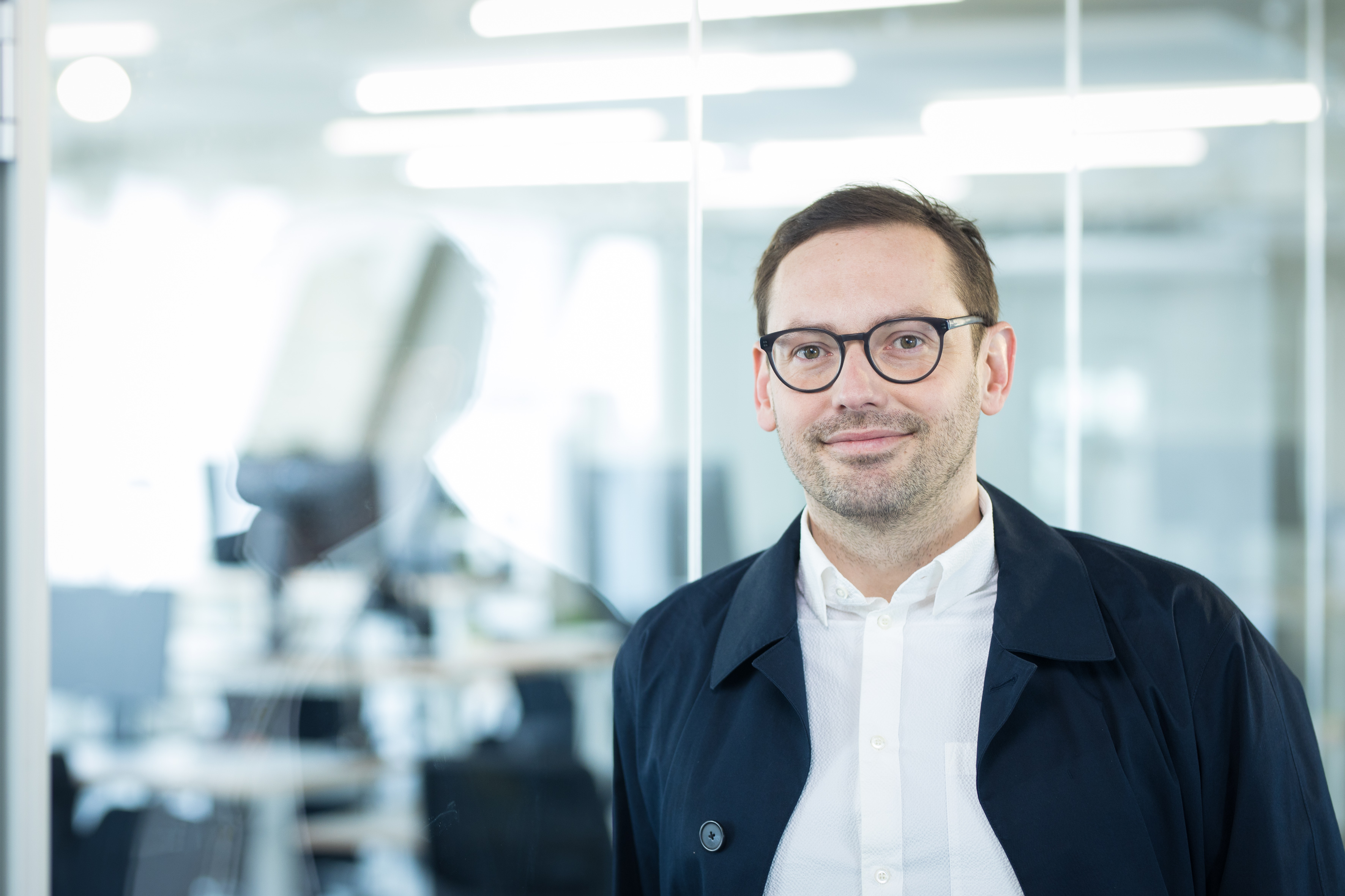 Martin Jordan, Head of Design beim DigitalService, steht in einem Bürogebäude vor einer Glaswand. Er trägt eine Brille, ein weißes Hemd und eine leichte blaue Jacke.