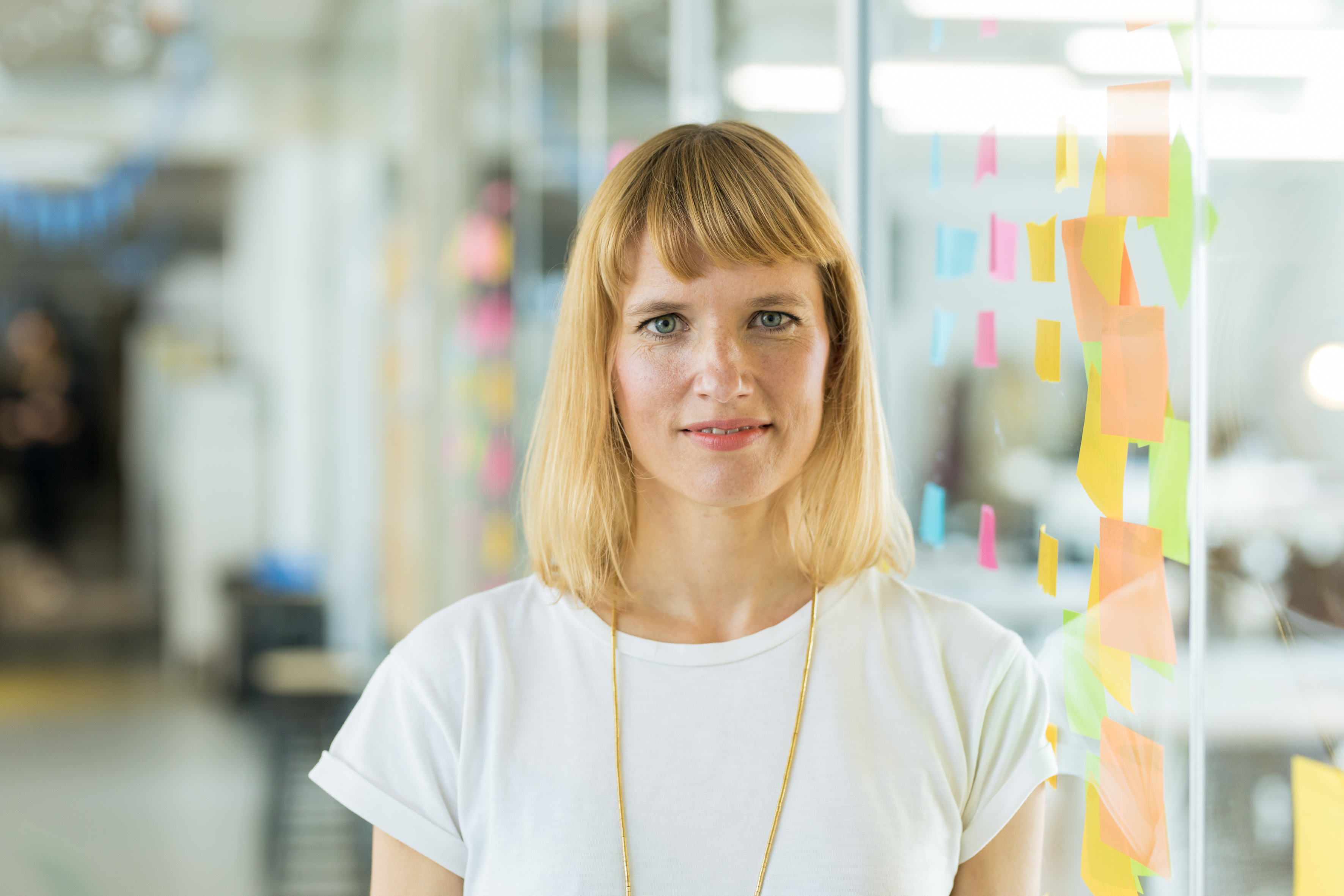UX Designerin Charlotte steht in einem Büro vor einer Glaswand mit Notizzetteln. Sie trägt ein weißes T-Shirt und eine goldene Kette