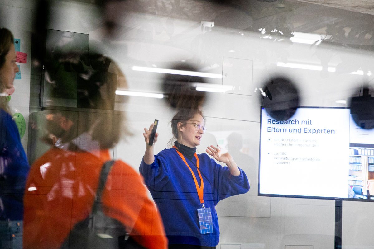 Eine Frau in einem blauen Pullover mit orangefarbenem Lanyard hält eine Präsentation in einem modernen Konferenzraum. Sie gestikuliert mit einer Hand und hält in der anderen ein Smartphone oder eine Fernbedienung. Hinter ihr befindet sich eine Leinwand mit einer Folie auf Deutsch, die den Titel „Research mit Eltern und Experten“ trägt.