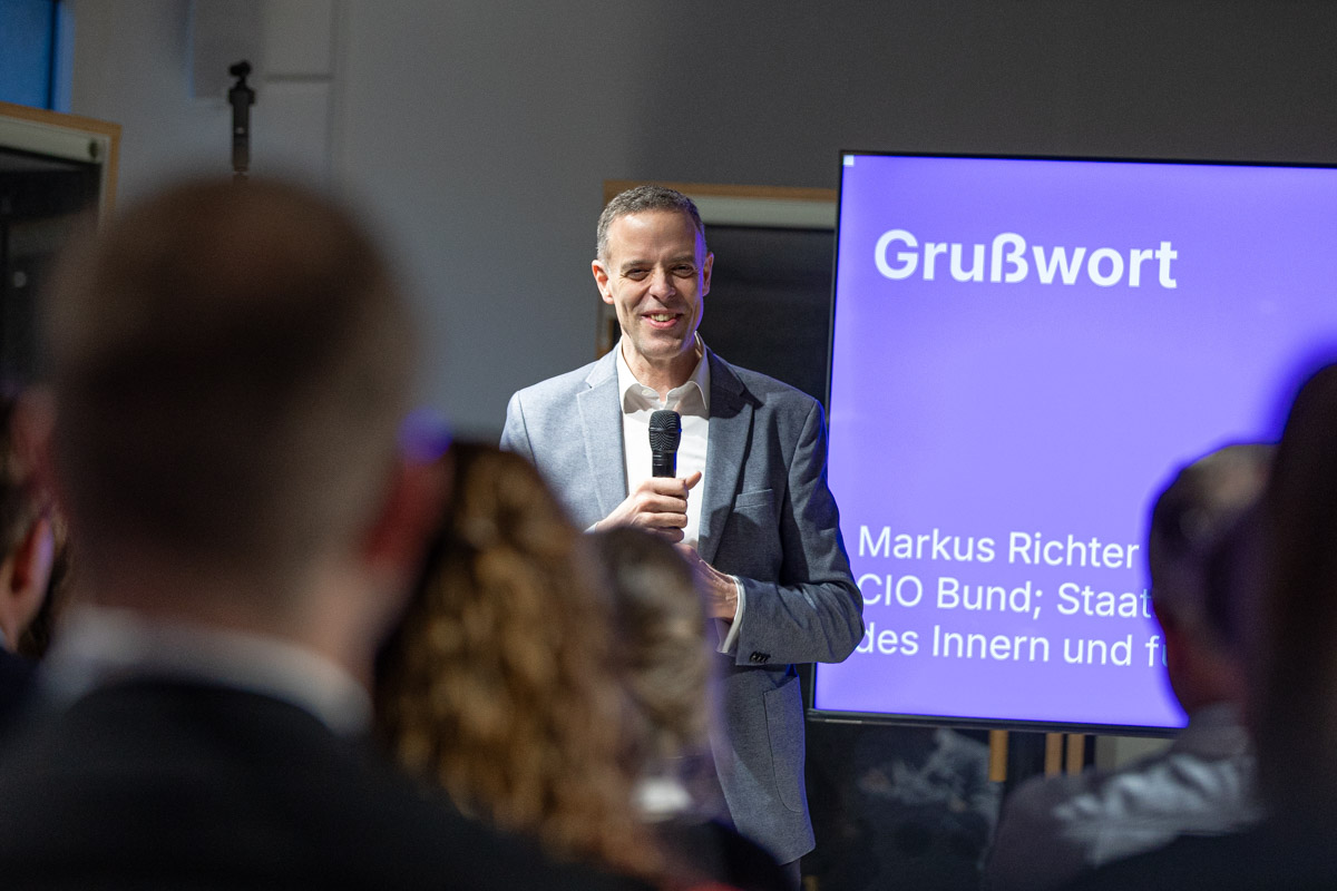 Ein Mann steht vor einem Bildschirm mit einem Mikrophon und lächelt. Auf dem Bildschirm steht „Grußwort“, „Markus Richter, CIO Bund“.