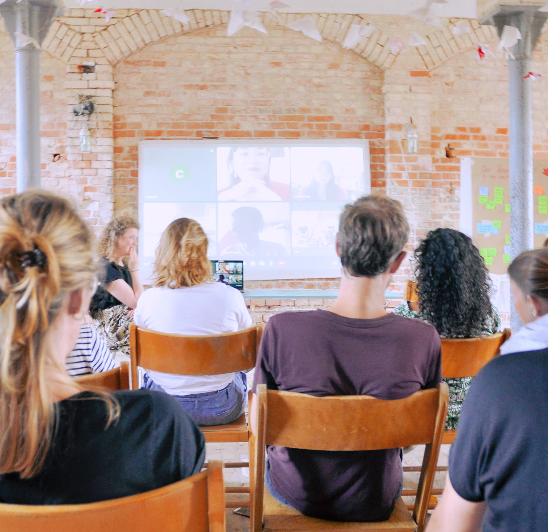 Several people sit together in a room during a hybrid meeting; the virtual participants can be seen on a screen