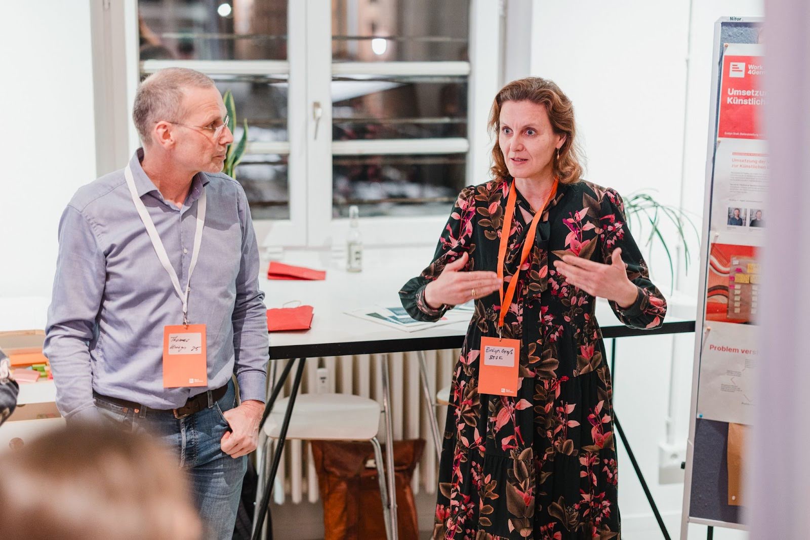 Zwei Personen stehen neben einer Plakatwand mit bunten Zetteln und präsentieren etwas.