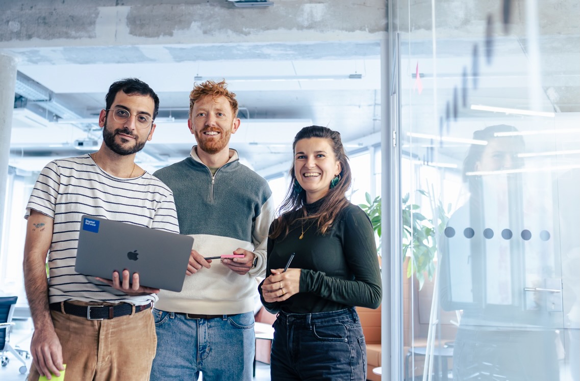 Drei Personen stehen in einem modernen Büro und lächeln in die Kamera. Einer hält einen Laptop, die anderen halten Stifte. Im Hintergrund sind Glaswände, Pflanzen und Büromöbel sichtbar.