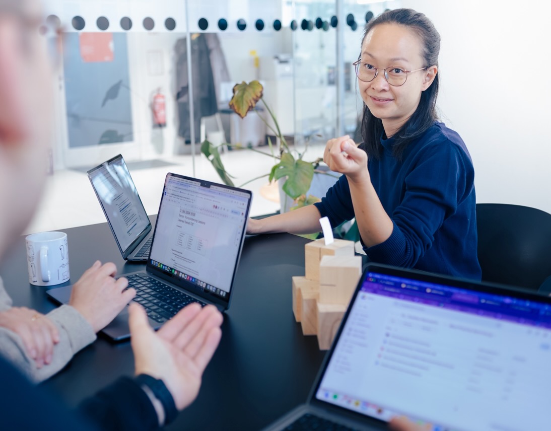 Eine Person sitzt an einem schwarzen Tisch und spricht mit zwei Personen. Vor ihr und den anderen stehen geöffnete Laptops mit sichtbaren Dokumenten und E-Mails auf den Bildschirmen. Auf dem Tisch liegen außerdem Holzklötze und eine Tasse mit Illustrationen.