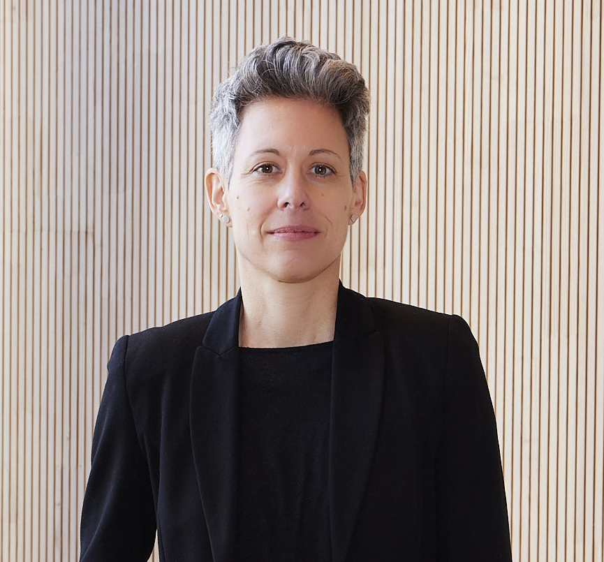 Julia Borggräfe stands in front of a light beige wall and looks into the camera with a slight smile; she wears a black blazer over a black top; her hair is short and graying