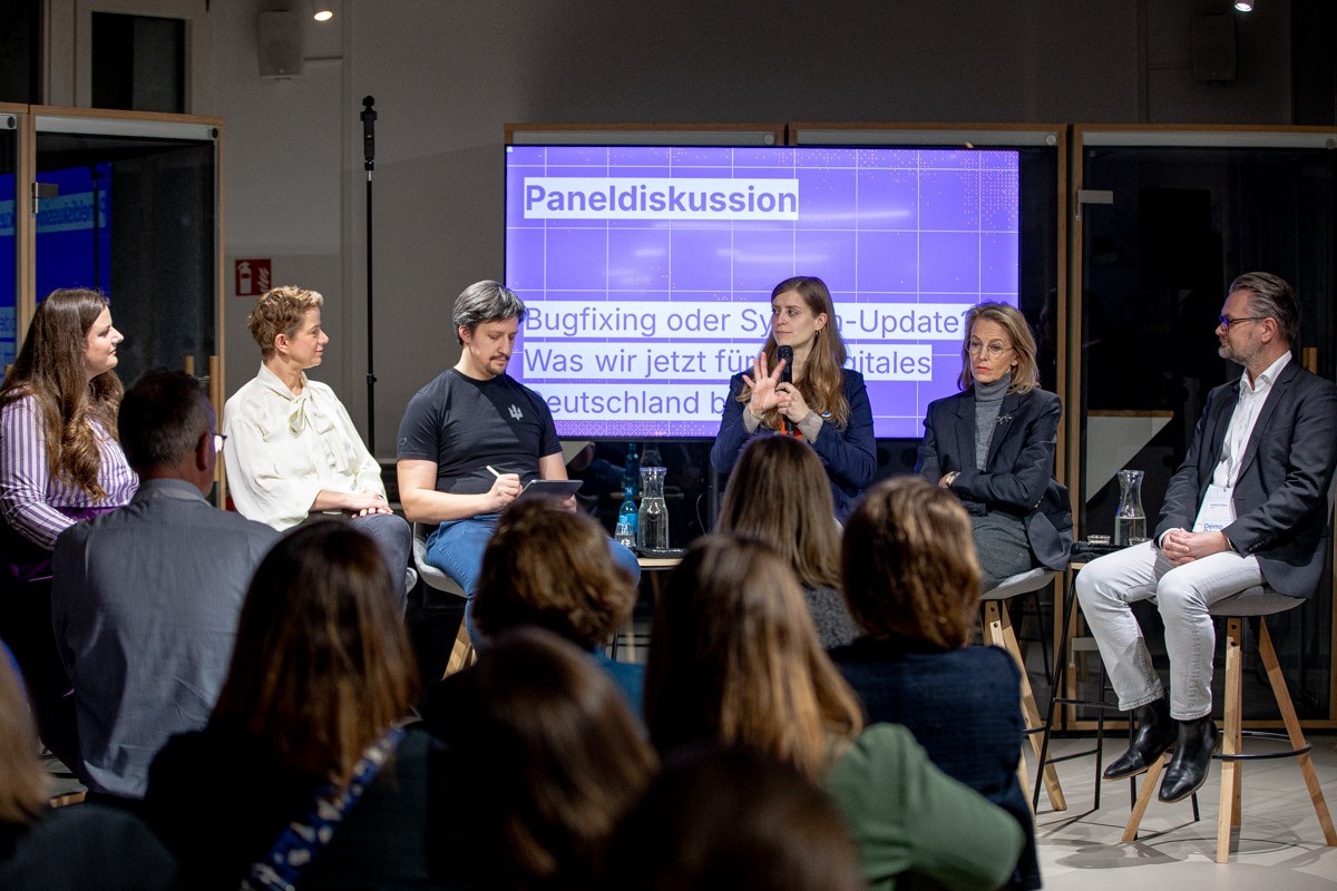 Eine Paneldiskussion mit fünf Teilnehmenden auf einer Bühne. Christina Lang spricht ins Mikrofon, während das Publikum zuhört. Im Hintergrund zeigt eine Leinwand den Titel der Diskussion „Bugfixing oder System-Update? Was wir jetzt für ein digitales Deutschland brauchen“