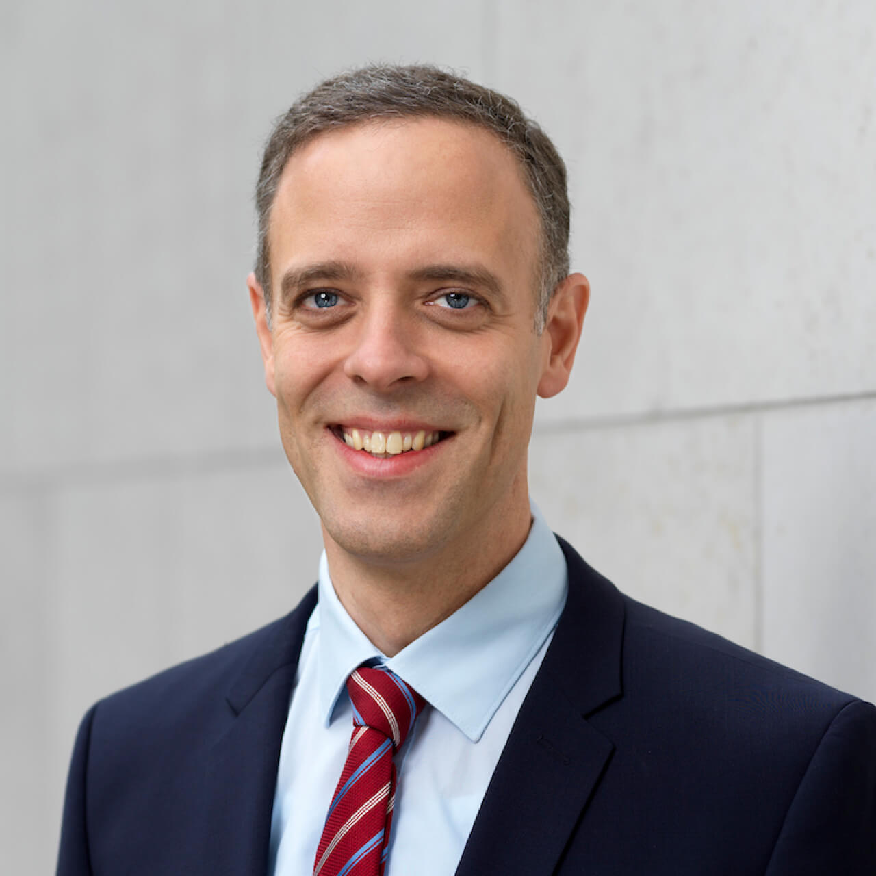 Supervisory Board member and federal CIO Markus Richter stands in front of a light gray stone wall and looks into the camera with a big smile showing his teeth; he wears a blue jacket, light blue shirt and dark red tie with blue and gold stripes; he has short, slightly graying hair