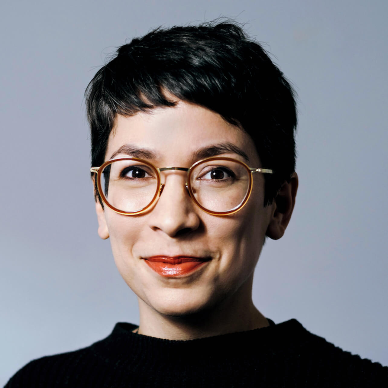 Supervisory Board member Julia Kloiber stands in front of a grey-blue background and smiles at the camera; she has short black hair and wears round golden-brown glasses and a black top; her lips are painted red