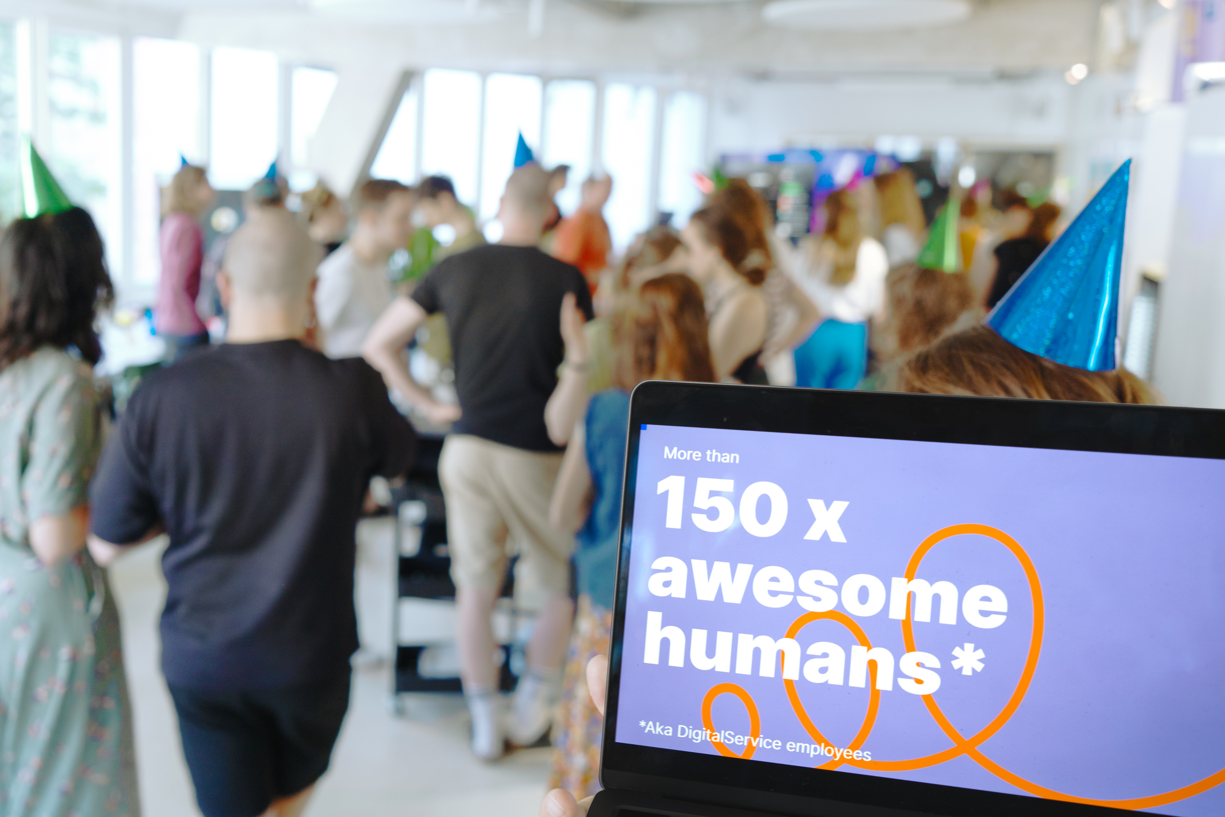 In the foreground, a laptop screen shows that more than 150 employees now work at DigitalService; in the background, numerous employees can be seen celebrating together in a blur
