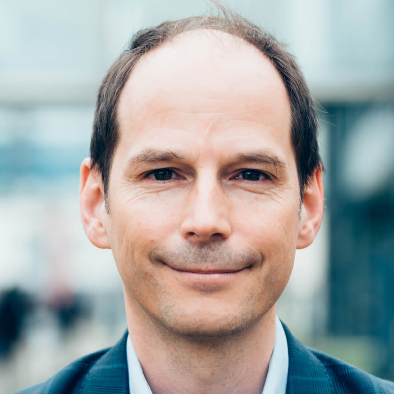 Supervisory Board member Peter Parycek looks straight into the camera with a slight smile; he has short brown hair that is thin on top of his head; he wears a blue jacket and a light blue shirt; in the background it can be seen dimly and very blurry that he is standing outdoors on a street