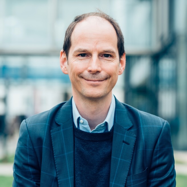 Aufsichtsratmitglied Peter Parycek blickt mit einem leichten Lächeln direkt in die Kamera; er hat kurze braune Haare, die am Oberkopf licht sind; er trägt ein blaues Sakko und ein hellblaues Hemd; im Hintergrund ist schemenhaft und sehr unscharf zu erkennen, dass er im Freien auf einer Straße steht