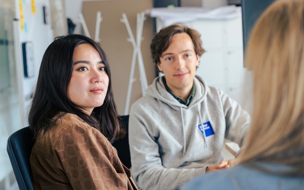 Zwei Mitarbeitende des DigitalService sitzen in einem Büro und schauen eine andere Person an, die nur von hinten zu sehen ist. Im Hintergrund sind ein Kleiderständer, Schränke und eine Pinnwand sichtbar.