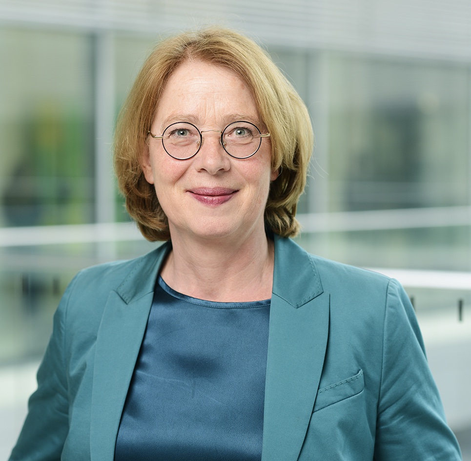 Tabea Rößner, member of the Bundestag, smiles slightly at the camera; she has shoulder-length strawberry blonde hair, wears round black glasses and a green blazer over a silky shimmering top; in the background a glass facade of an office building can be seen