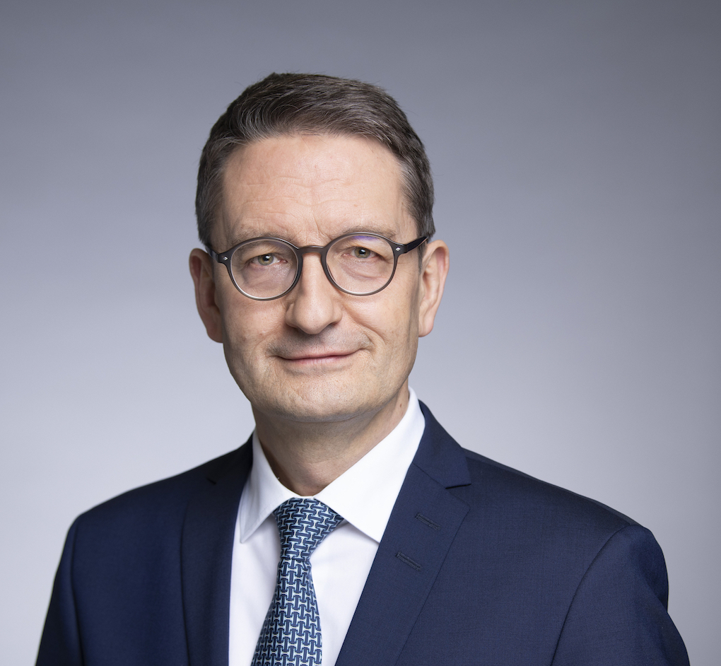 Stefan Schnorr, State Secretary in the Federal Ministry for Digital Affairs and Transport, is wearing a blue suit, white shirt and blue checked tie in front of a light gray background and smiles slightly at the camera; he has short brown hair and wears round charcoal gray glasses