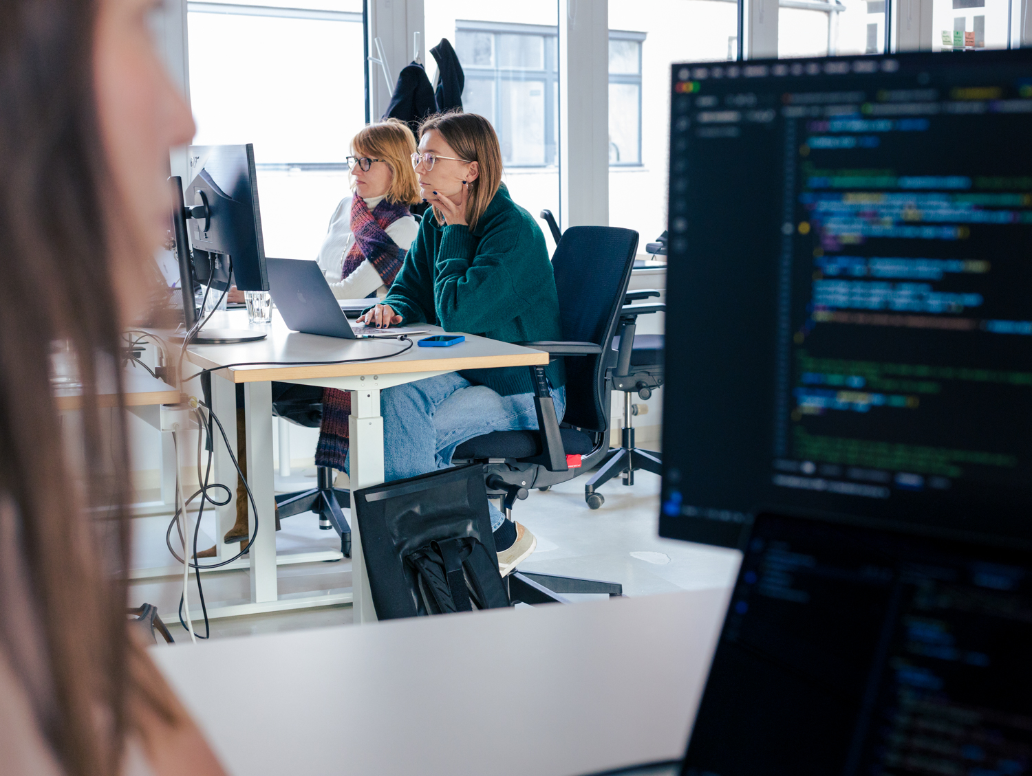 Im Büro des DigitalService ist auf einem Bildschirm im Vordergrund Software-Code zu sehen; im Hintergrund sitzen zwei Mitarbeitende an einem Tisch und arbeiten