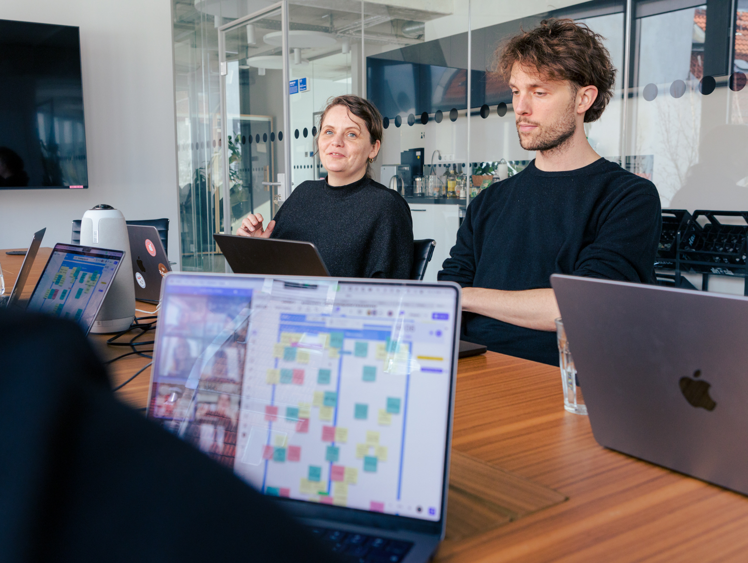 DigitalService employees sit at a long table with their laptops open and discuss; virtual participants can be seen on the laptops, along with a board with sticky notes.